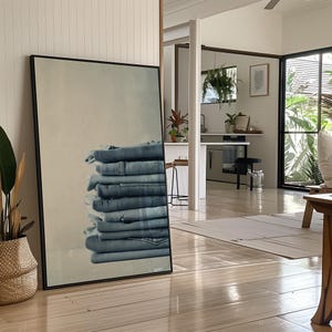 May include: A large framed print featuring a stack of folded blue denim jeans. The jeans are neatly arranged, showcasing their various shades of blue. The print is displayed in a bright, airy room with natural light and wooden floors.