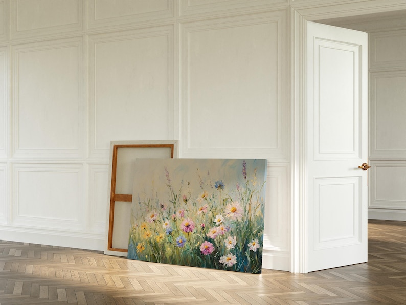 May include: An oil painting on canvas depicts a field of wildflowers in shades of pink, yellow, and blue. The artwork is leaning against a wall in a room with white paneling and a wooden floor. A white door is slightly ajar.