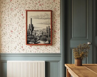 May include: A framed black and white desert landscape art print featuring cacti and a cloudy sky. The artwork is displayed on a floral wallpapered wall with a red frame. The room has a blue door and a wooden table.