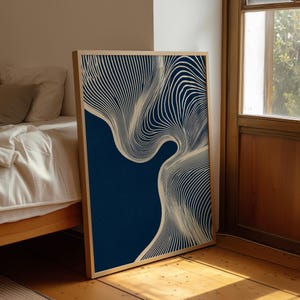 May include: A framed abstract art print featuring a navy blue background with flowing, cream-colored lines. The artwork is displayed in a light wood frame, leaning against a wall near a window and bed.