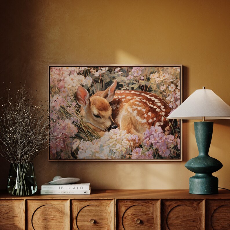 May include: A framed art print featuring a fawn resting amidst a bed of flowers. The fawn has brown fur with white spots and is surrounded by white and pink blossoms. The artwork is displayed above a wooden cabinet, with a lamp and vase.
