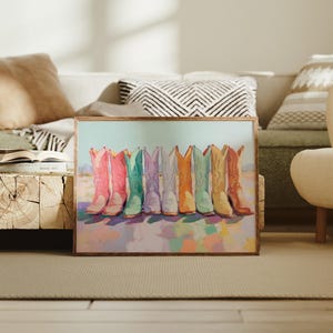 May include: A framed art print featuring a row of colorful cowboy boots. The boots are painted in shades of pink, green, purple, orange, and yellow. The background is a soft, pastel landscape. The artwork is displayed in a living room setting.