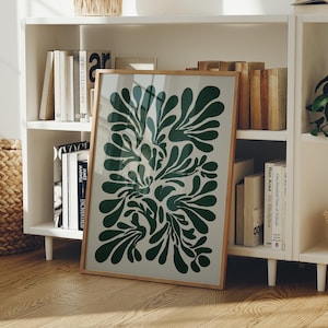 May include: A framed art print with a green abstract botanical design on a light beige background. The artwork is displayed on a wooden floor, leaning against a white bookshelf filled with books. The frame is a light wood colour.