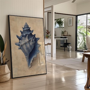 May include: A framed print featuring a detailed illustration of a large, blue seashell. The artwork is set against a beige background and framed in black. The print leans against a wall in a well-lit room with wooden floors, suggesting a coastal or nautical theme.