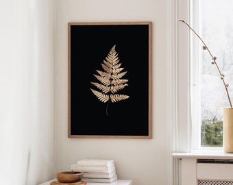 May include: A framed art print featuring a gold fern leaf against a black background. The fern is detailed with intricate veining, set within a natural wood frame. The artwork is displayed in a bright room with a white table and window.