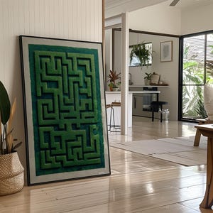 Puede incluir: Una obra de arte enmarcada con un diseño de laberinto verde sobre un fondo verde claro. El laberinto, con sus caminos interconectados, está enmarcado en un marco negro. La obra se apoya en una pared en una habitación luminosa con suelo de madera.