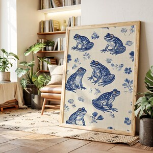 May include: A framed print featuring a repeating pattern of blue frogs and floral elements on a beige background. The artwork has a light wooden frame and is displayed in a well-lit room with plants and books. The print's design is reminiscent of vintage botanical illustrations.