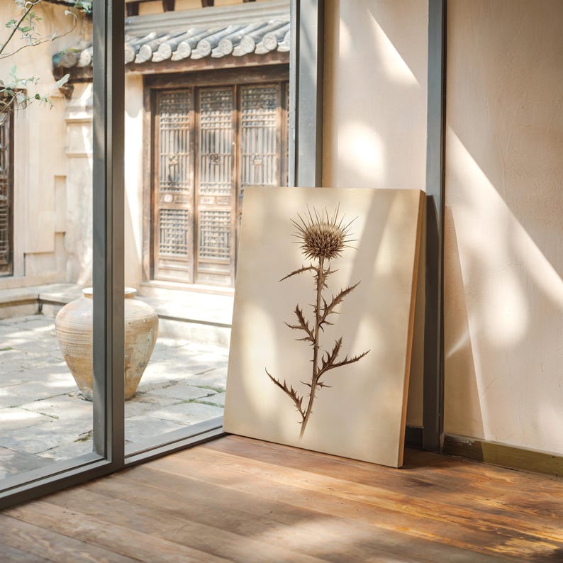 May include: A framed botanical print of a thistle plant. The artwork features a detailed illustration of the plant's spiky flower head and stem, rendered in shades of brown and beige. The print is leaning against a wall near a window.