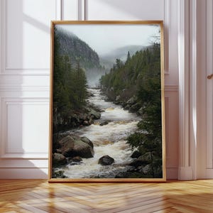 May include: A framed landscape print featuring a river flowing through a valley. The artwork showcases a misty mountain scene with lush green trees and large rocks along the riverbanks. The frame is a warm, natural wood tone.