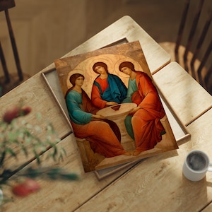 May include: An icon depicting three figures in robes, seated around a table. The figures have halos and are rendered in warm tones of red, blue, and orange. The icon is in a wooden frame, resting on a wooden table.
