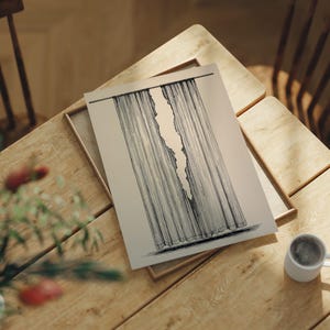 Puede incluir: Una impresión artística en blanco y negro que representa una cortina rasgada. La obra de arte está sobre un fondo beige claro y se presenta en una bandeja de madera. Una taza de café está sobre una mesa de madera.