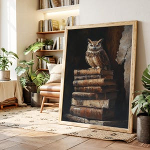 Op de afbeelding: Een ingelijste print met een gedetailleerd schilderij van een bruine uil die bovenop een stapel oude boeken zit. Het kunstwerk heeft een donkere, sfeervolle esthetiek met de nadruk op de kenmerken van de uil en de textuur van de boeken. De lijst is van licht hout.