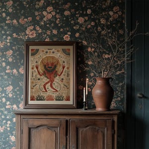 May include: A framed artwork featuring a red demon-like figure with horns and outstretched arms. The artwork is displayed on a wooden cabinet, with a vase of branches and lit candles nearby. The background is a floral wallpaper.