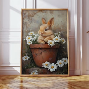 May include: A framed art print featuring a charming, light brown bunny nestled in a terracotta pot filled with white daisies. The bunny has long ears and a sweet expression. The artwork has a soft, painterly style with a neutral background.