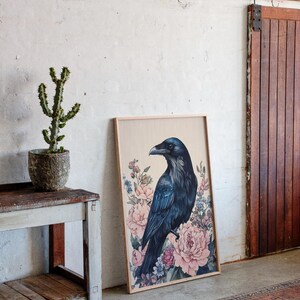 May include: A framed art print featuring a dark blue raven perched amidst pink and blue floral elements. The artwork is displayed against a neutral background, leaning against a wall near a wooden door and a small table with a potted cactus.