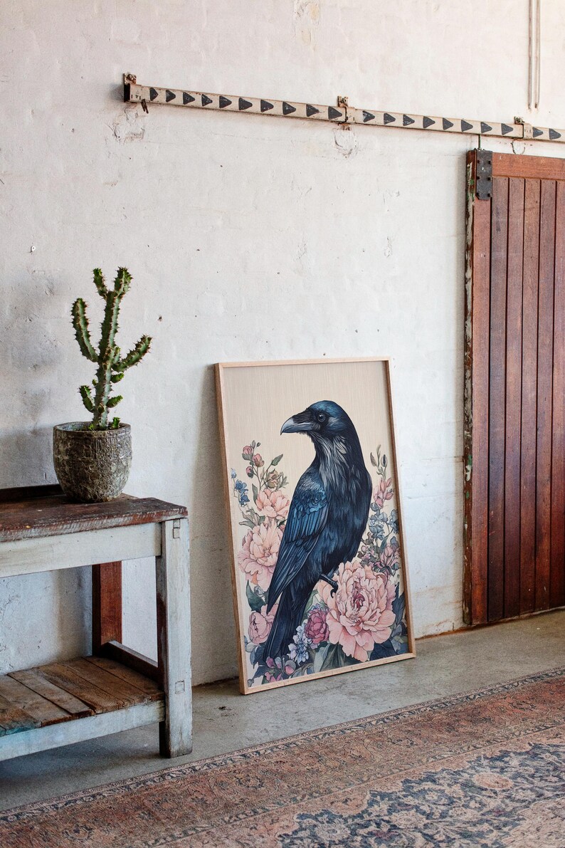 May include: A framed art print featuring a dark blue raven perched amidst pink and blue floral elements. The artwork is displayed against a neutral background, leaning against a wall near a wooden door and a small table with a potted cactus.