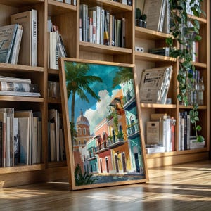 May include: A framed watercolor print depicting a colorful street scene with buildings in shades of pink, turquoise, and yellow. Palm trees and a domed building are also visible. The artwork is displayed on a wooden floor in front of a bookshelf.