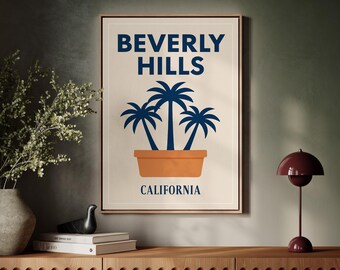 May include: Framed art print featuring the words "BEVERLY HILLS" in navy blue above a graphic of three palm trees in an orange pot, with "CALIFORNIA" below. The artwork is displayed on a wooden cabinet.