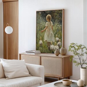 May include: A painting of a young girl in a white dress walking through a field with a flock of white ducks. The painting is framed in a light wood frame and is hanging on a white wall.