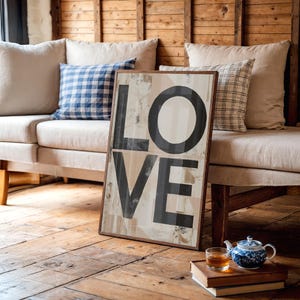 May include: A framed wooden sign with the word "LOVE" in large black letters. The background has a neutral color palette. The sign is leaning against a sofa with pillows, and a teapot and teacup are on the floor.