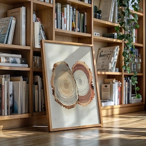 May include: A framed artwork featuring two stylized cross-sections of tree trunks. The artwork displays concentric rings in shades of brown, beige, and gray, set against a white background. The frame is made of light-colored wood, and the artwork is leaning against a bookshelf.
