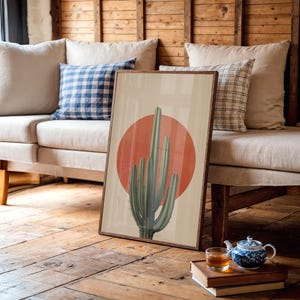 May include: A framed art print depicting a green cactus against a red sun on a beige backdrop. The artwork is displayed on a wooden floor, leaning against a beige sofa with blue and white checked cushions.