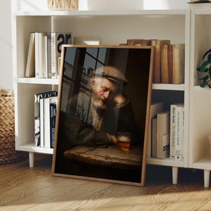 May include: A framed print of an older man with a beard, wearing a cap, resting his head on his hand at a wooden table. A glass of amber liquid sits on the table. The print is displayed on a white bookshelf with books.