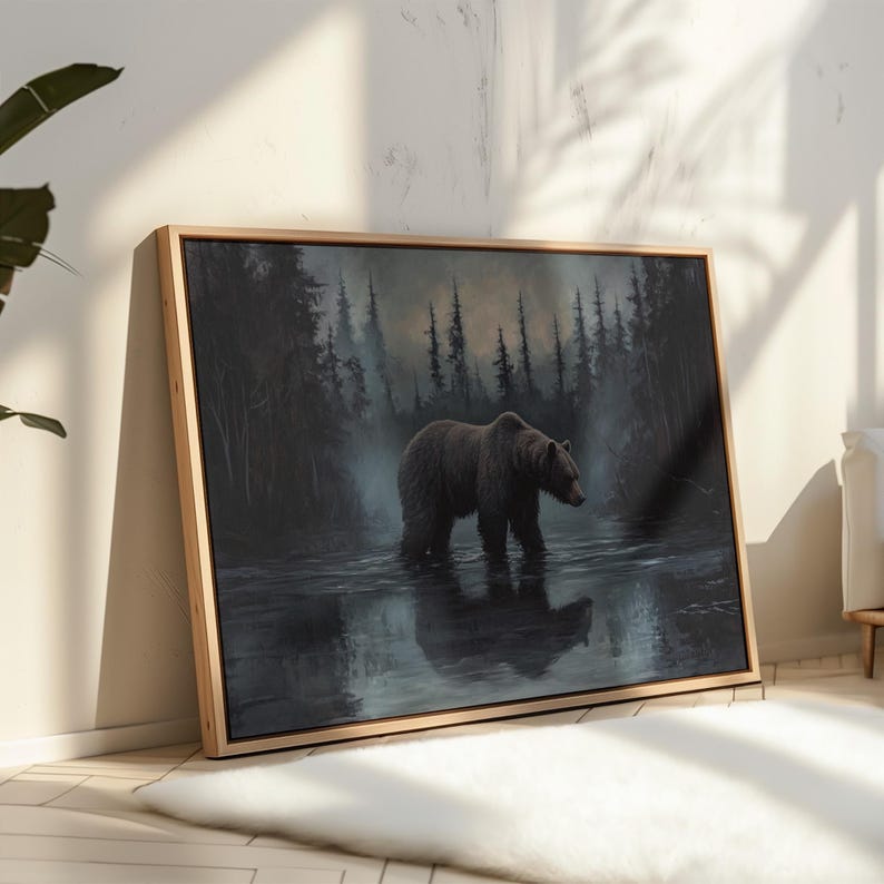 Op de afbeelding: Een ingelijste canvasprint toont een bruine beer die door een donkere, mistige rivier waadt. De beer is het middelpunt, met een bos op de achtergrond en zijn weerspiegeling in het water. Het kunstwerk is in een licht houten frame.