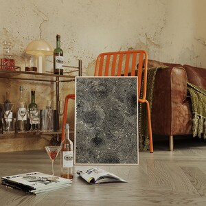 May include: A framed abstract art print featuring a pattern of black concentric circles. The artwork is displayed near a bar cart with bottles, glasses, and a cocktail. A bottle of wine and a magazine are on the floor. An orange chair and a brown leather sofa are in the background.