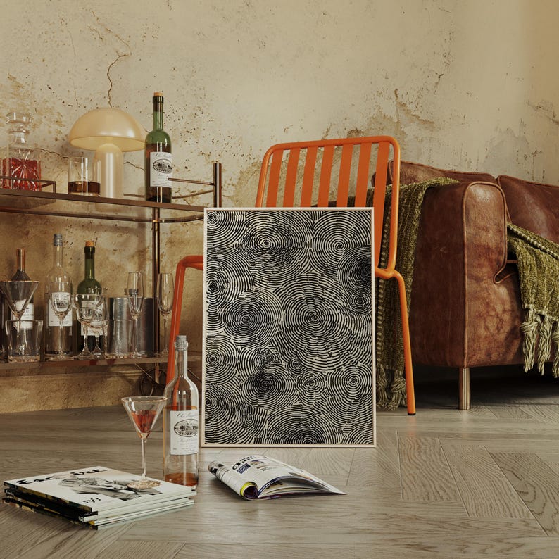 May include: A framed abstract art print featuring a pattern of black concentric circles. The artwork is displayed near a bar cart with bottles, glasses, and a cocktail. A bottle of wine and a magazine are on the floor. An orange chair and a brown leather sofa are in the background.