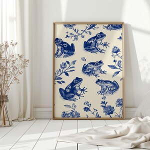 May include: A framed art print featuring a repeating pattern of blue frogs and floral elements on a cream background. The artwork is displayed in a light-colored wooden frame, leaning against a white wall.