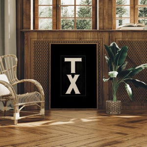 May include: A framed black and white print featuring the letters "TX" in a bold, serif font. The artwork is displayed in a room with wooden paneling, a wicker chair, and a potted plant, creating a rustic and inviting atmosphere. The print is a decorative wall art.