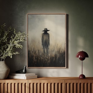 Peut inclure: Une &oelig;uvre encadr&eacute;e repr&eacute;sente une silhouette solitaire portant un chapeau &agrave; larges bords dans un champ d'herbes hautes. La peinture utilise une palette de couleurs sourdes de bruns, de gris et de beige, avec un cadre sombre. Une lampe de table bordeaux et des objets d&eacute;coratifs sont pos&eacute;s sur un meuble en bois.