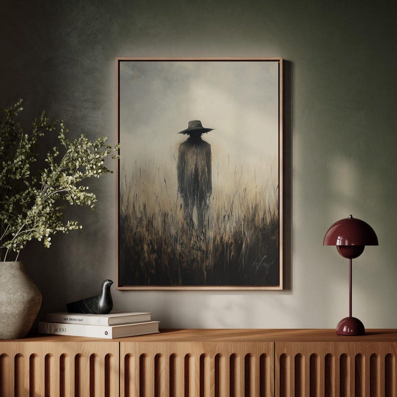 Peut inclure: Une &oelig;uvre encadr&eacute;e repr&eacute;sente une silhouette solitaire portant un chapeau &agrave; larges bords dans un champ d'herbes hautes. La peinture utilise une palette de couleurs sourdes de bruns, de gris et de beige, avec un cadre sombre. Une lampe de table bordeaux et des objets d&eacute;coratifs sont pos&eacute;s sur un meuble en bois.