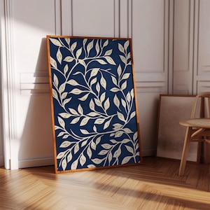 May include: A framed art print featuring a navy blue background with a cream-colored leaf pattern. The artwork is encased in a wooden frame and leans against a white wall. The print's design is a botanical theme.