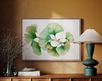 May include: A framed watercolor painting of ginkgo leaves in shades of green and white. The artwork is displayed on a wooden cabinet, next to a teal lamp and a vase with dried branches. The painting has a natural, botanical theme.