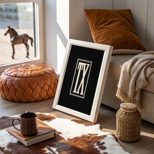 May include: A framed artwork with the letters "TX" in a serif font against a black background. The white frame leans against a wall, near a brown leather ottoman, a cowhide rug, and a brown mug on a book.