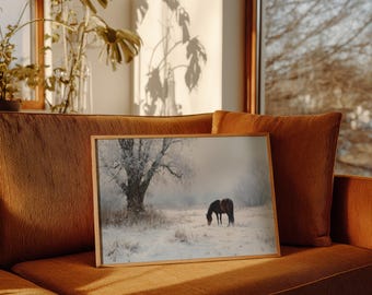 May include: A framed landscape print featuring two brown horses grazing in a snowy field. A large tree is visible on the left side of the image, with a soft, muted color palette. The print is displayed on a rust-colored couch.