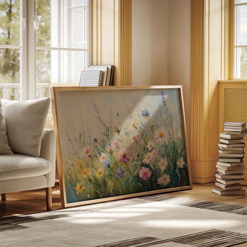 May include: A framed floral painting featuring a meadow of colorful wildflowers in shades of pink, yellow, blue, and white. The artwork is displayed in a light-toned wooden frame, leaning against a wall in a well-lit room with a neutral color palette.