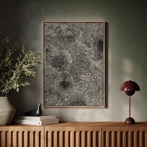 May include: A framed abstract art print featuring a pattern of black concentric circles on a cream background. The artwork hangs above a wooden cabinet, with a decorative bird, books, and a burgundy table lamp.