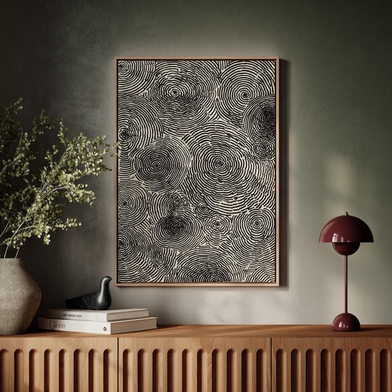 May include: A framed abstract art print featuring a pattern of black concentric circles on a cream background. The artwork hangs above a wooden cabinet, with a decorative bird, books, and a burgundy table lamp.