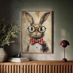 May include: A painting of a brown rabbit wearing a red and white checkered bow tie and round glasses. The rabbit is looking directly at the viewer. The painting is framed in a light wood frame.