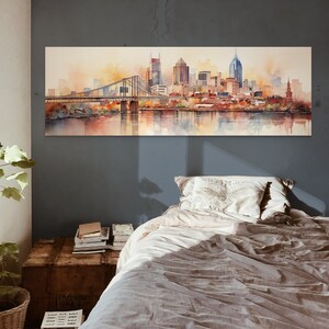Op de afbeelding: Een aquarel schilderij van een skyline met een brug op de voorgrond. De stad is afgebeeld in tinten bruin, oranje en geel, met een rivier op de voorgrond. Het schilderij is ingelijst en hangt aan een muur boven een bed.