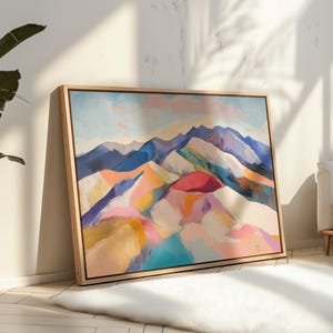 May include: Abstract mountain landscape painting in a wooden frame. The artwork features a colourful palette of blues, purples, pinks, yellows, and oranges, depicting a stylised mountain range under a soft, pastel sky. The painting is leaning against a white wall.
