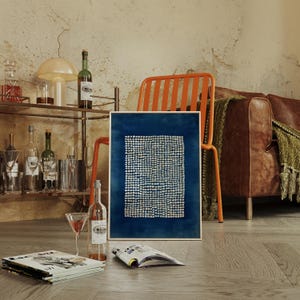 May include: A framed abstract art print with a dark blue background and a pattern of small white dots. The artwork is displayed in a room with a brown leather sofa, orange chair, and a bar cart with bottles and glasses.