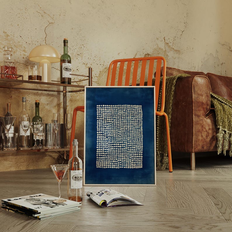 May include: A framed abstract art print with a dark blue background and a pattern of small white dots. The artwork is displayed in a room with a brown leather sofa, orange chair, and a bar cart with bottles and glasses.