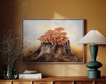 Op de afbeelding: Een ingelijst schilderij toont een cluster van rood-wit gestippelde paddenstoelen op een verweerde boomstronk. Het kunstwerk is geplaatst tegen een achtergrond van een veld en een bewolkte lucht. Het schilderij wordt tentoongesteld op een houten kast, naast een groene lamp.