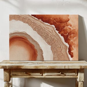 Può includere: Stampa artistica su tela rettangolare con un design astratto nei toni del marrone, beige e bianco. L'opera d'arte assomiglia a una sezione trasversale di una geode o a una mappa topografica, con texture a strati e dettagli di linee intricati. La stampa è esposta su un tavolo di legno.