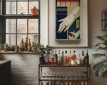 May include: A framed artwork depicting a vintage matchbox with a hand illustration and Japanese text. The matchbox contains red-tipped matches. A bar cart with various bottles of liquor and glassware is in the foreground. A window with plants is on the left.
