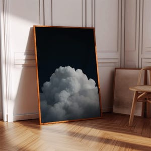 Op de afbeelding: Een ingelijste kunstdruk met een dramatische wolkenformatie tegen een donkerblauwe achtergrond. De wolk is een pluizige witte massa, en de lijst is van warm hout. Het kunstwerk staat tegen een witte muur.